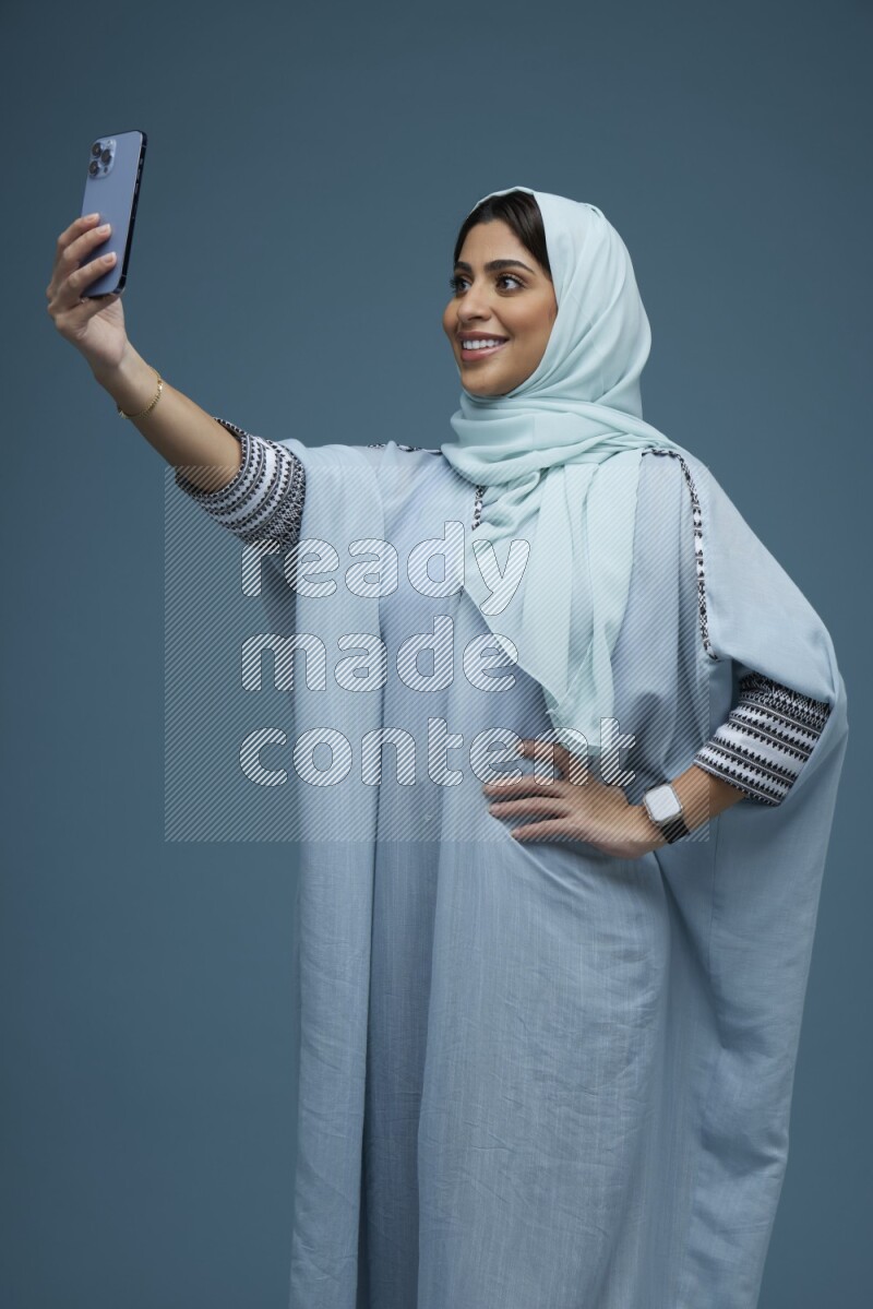 A Saudi woman Taking a Selfie on a blue background wearing a blue Abaya with hijab