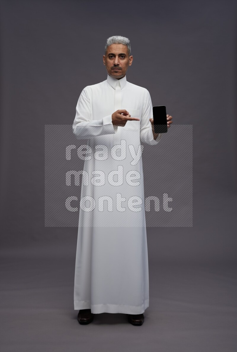 Saudi man wearing thob standing showing phone to camera on gray background