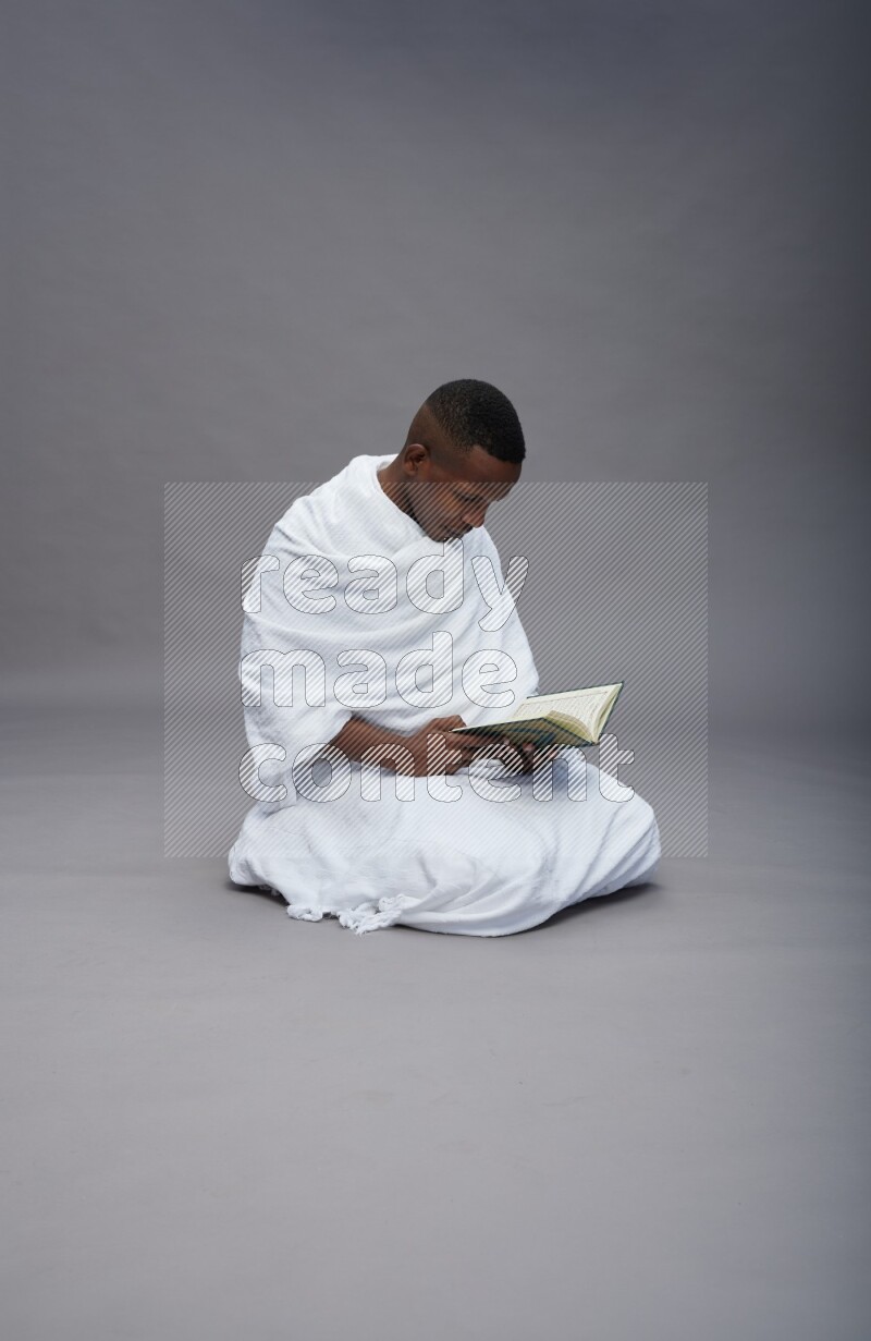 A man wearing Ehram sitting on floor reading quran on gray background