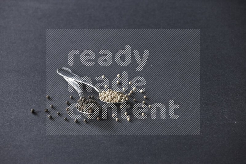 ملعقتان معدنيتان ممتلئتان بحبوب الفلفل الأسود والأبيض مع حبوب متناثرة على أرضية سوداء