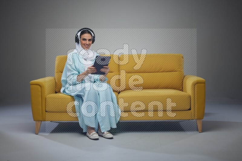 A Saudi woman wearing a light blue Abaya and a white head scarf sitting on a yellow sofa and holding an iPad while wearing headphone eye level on a grey background