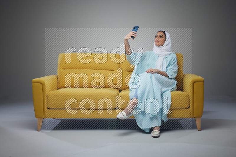 A Saudi woman wearing a light blue Abaya and white head scarf sitting on a yellow sofa and taking selfies with her phone eye level on a grey background