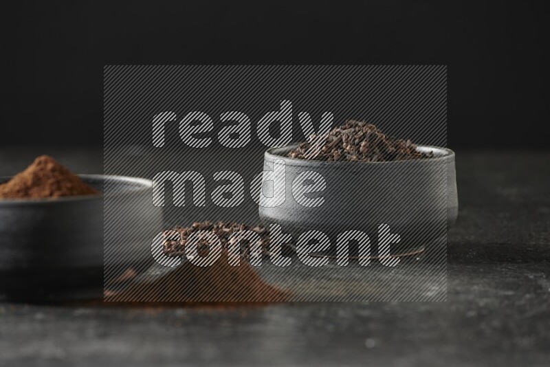 2 Black pottery bowls full of cloves and the other full of cloves powder on textured black flooring
