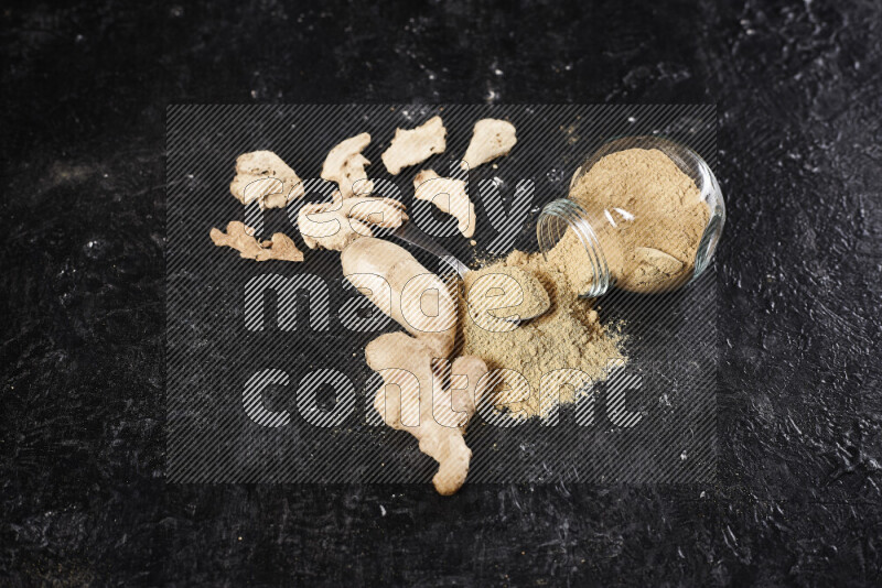 A glass jar full of ground ginger powder flipped with some spilling powder on black background