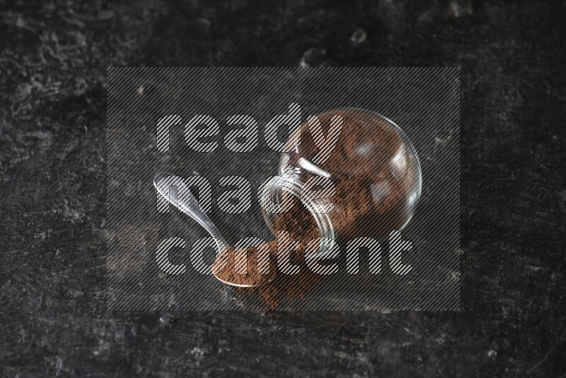 وعاء زجاجي للبهارات مقلوب وملعقة معدنية ممتلئان ببودرة القرنفل على أرضية سوداء