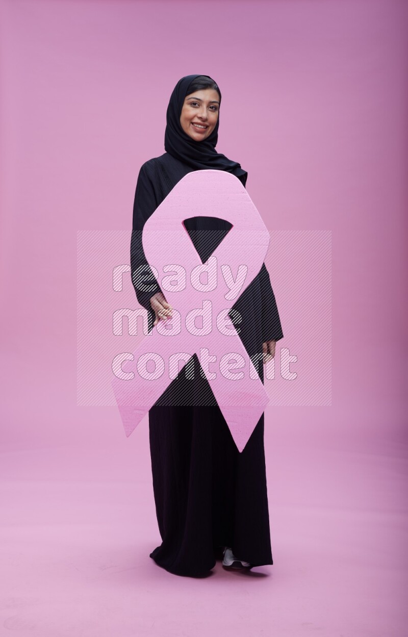 Saudi woman wearing pink ribbon on abaya standing holding awareness ribbon on plain pink background