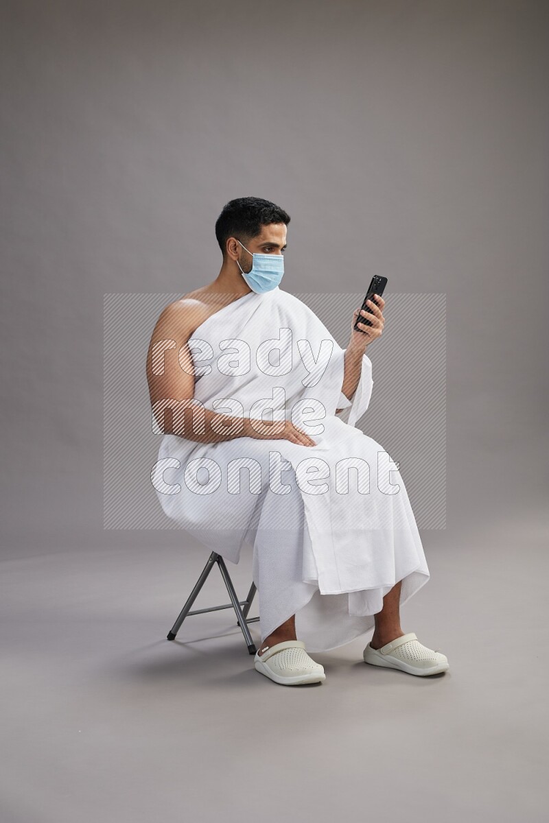 A man wearing Ehram with face mask sitting on chair texting on phone on gray background