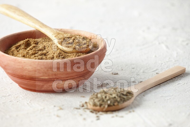 A wooden bowl and 2 wooden spoons full of cumin powder and cumin seeds on textured white flooring