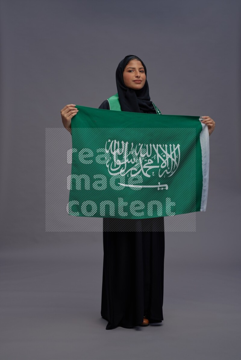 Saudi woman wearing Abaya standing holding Saudi flag on gray background