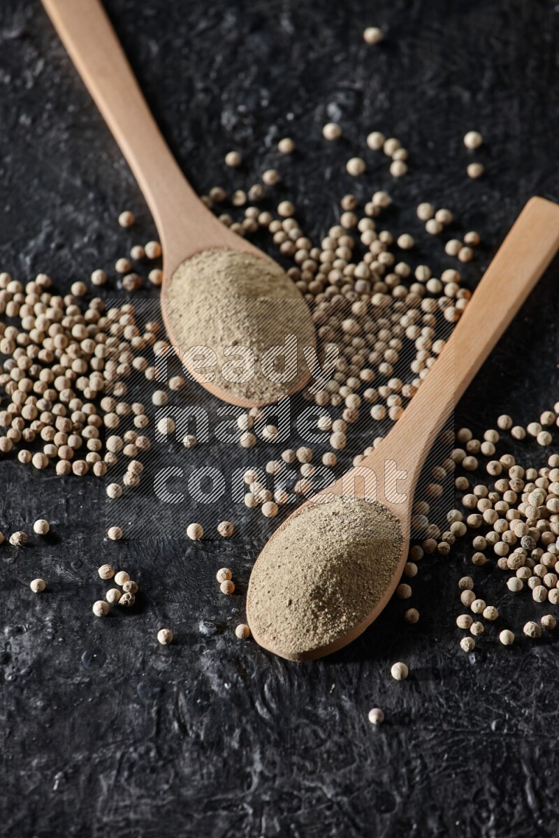 2 wooden spoons full of white pepper powder with white pepper beads on textured black flooring