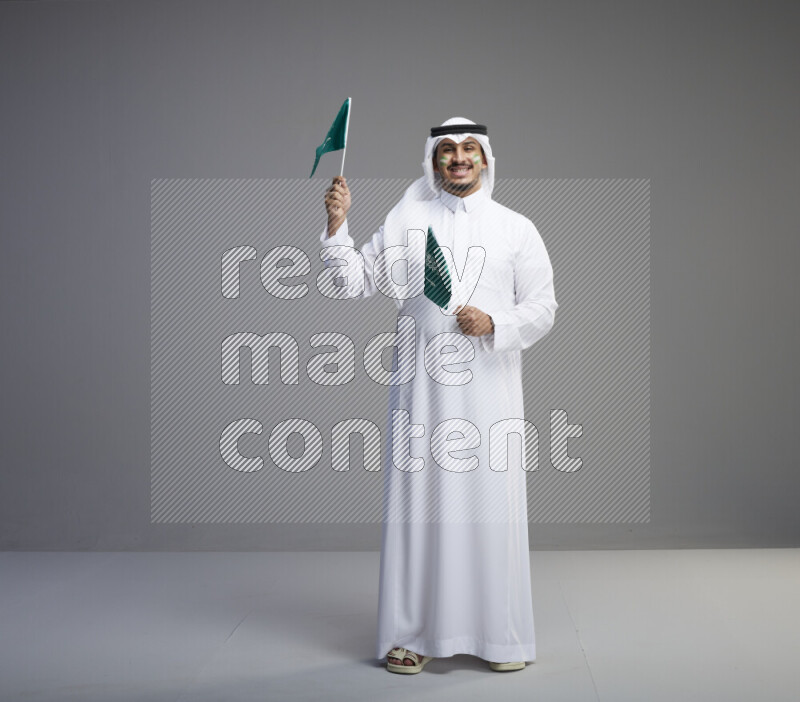 A Saudi man standing wearing thob and white shomag with face painting raising small saudi flag on gray background