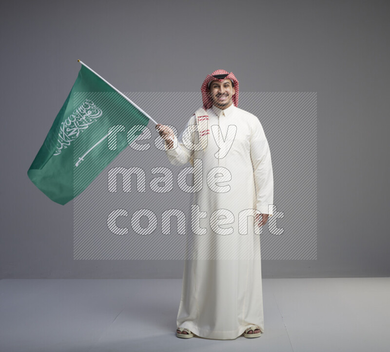 A Saudi man standing wearing thob and red shomag raising big Saudi flag on gray background