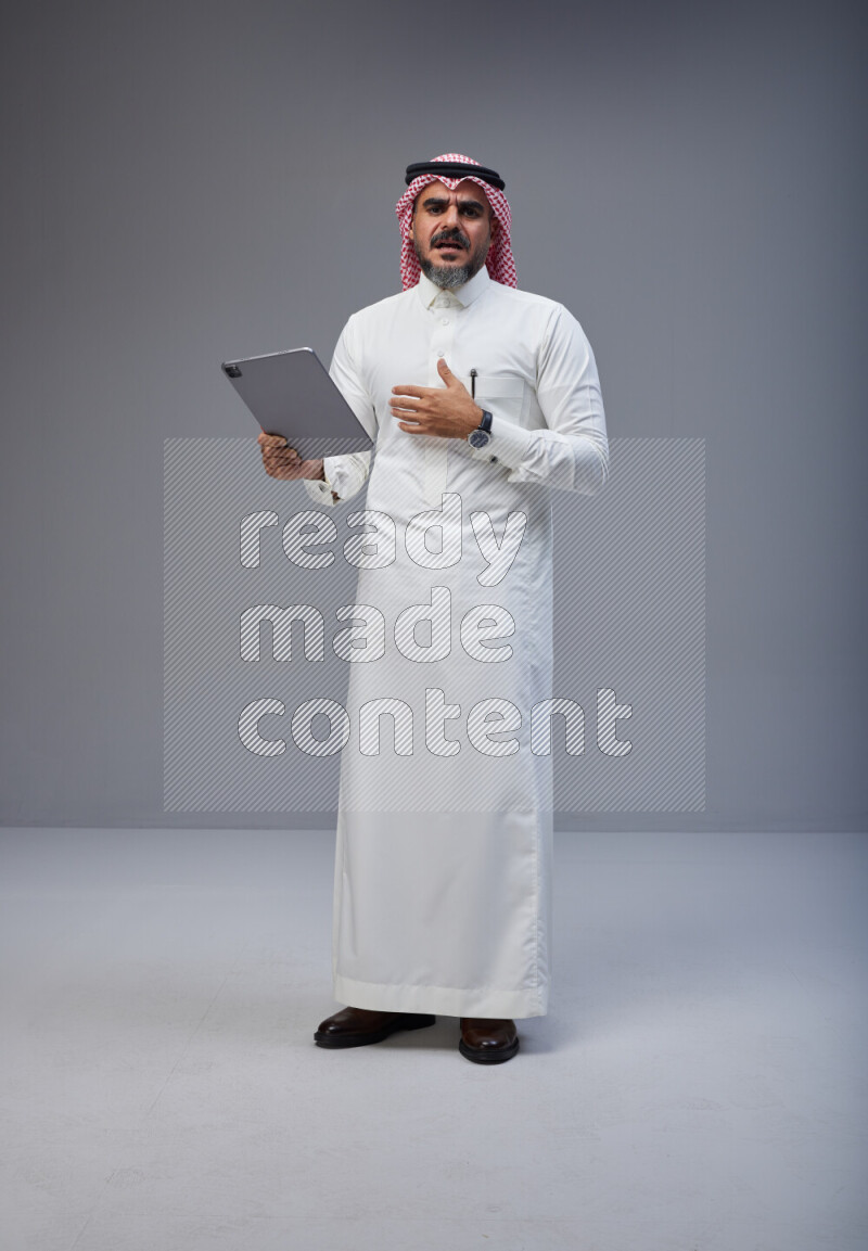 Saudi man Wearing Thob and red Shomag standing working on tablet on Gray background