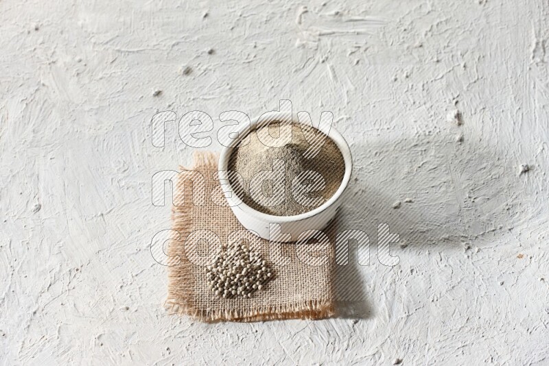 White pottery bowl full of white pepper powder set on a burlap piece of fabric with pepper beads and wooden pepper grinder on textured white flooring