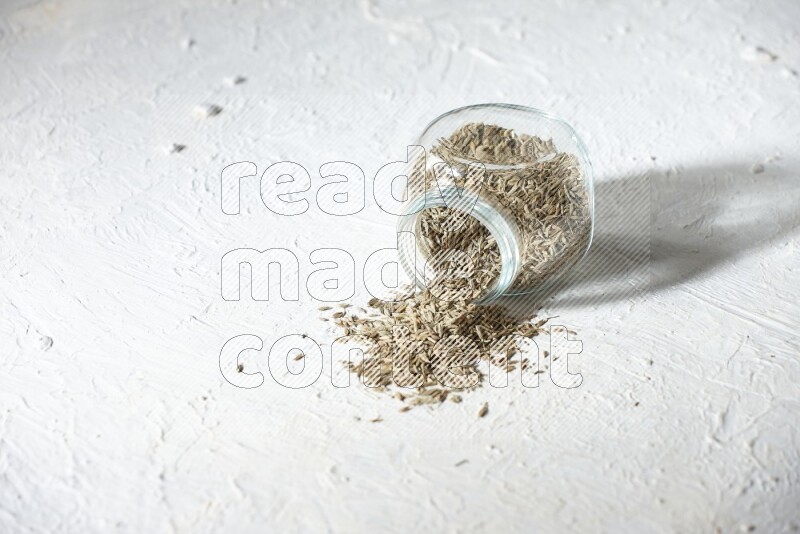 A flipped glass spice jar full of cumin seeds and the seeds spilled out on textured white flooring