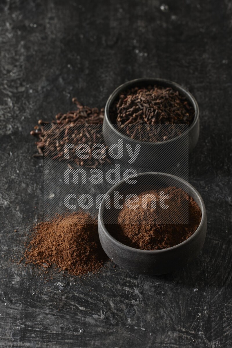 2 Black pottery bowls full of cloves and the other full of cloves powder on textured black flooring