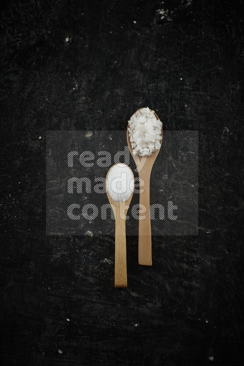 2 wooden spoons filled with fine and coarse salt on black background