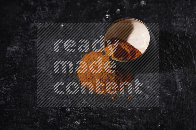 A black pottery bowl full of ground paprika powder with fallen powder from it on black background