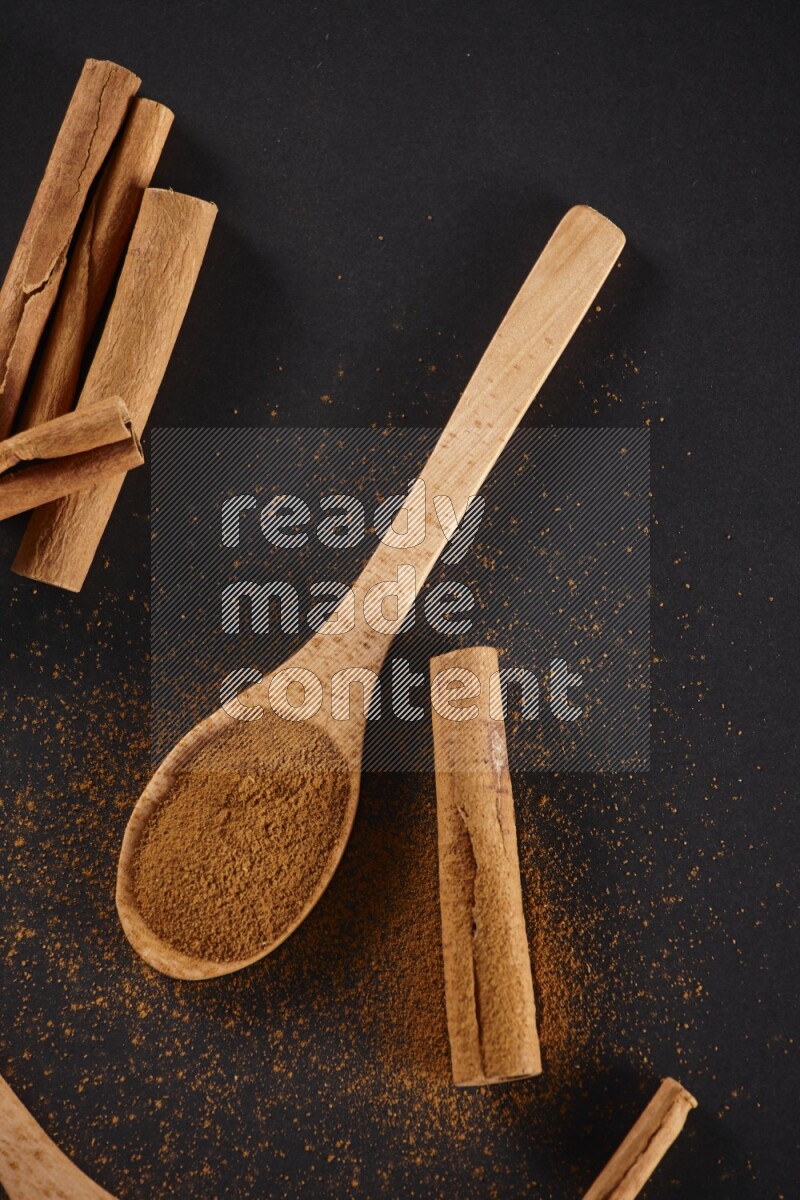 Cinnamon powder in two wooden spoons with cinnamon sticks on black background