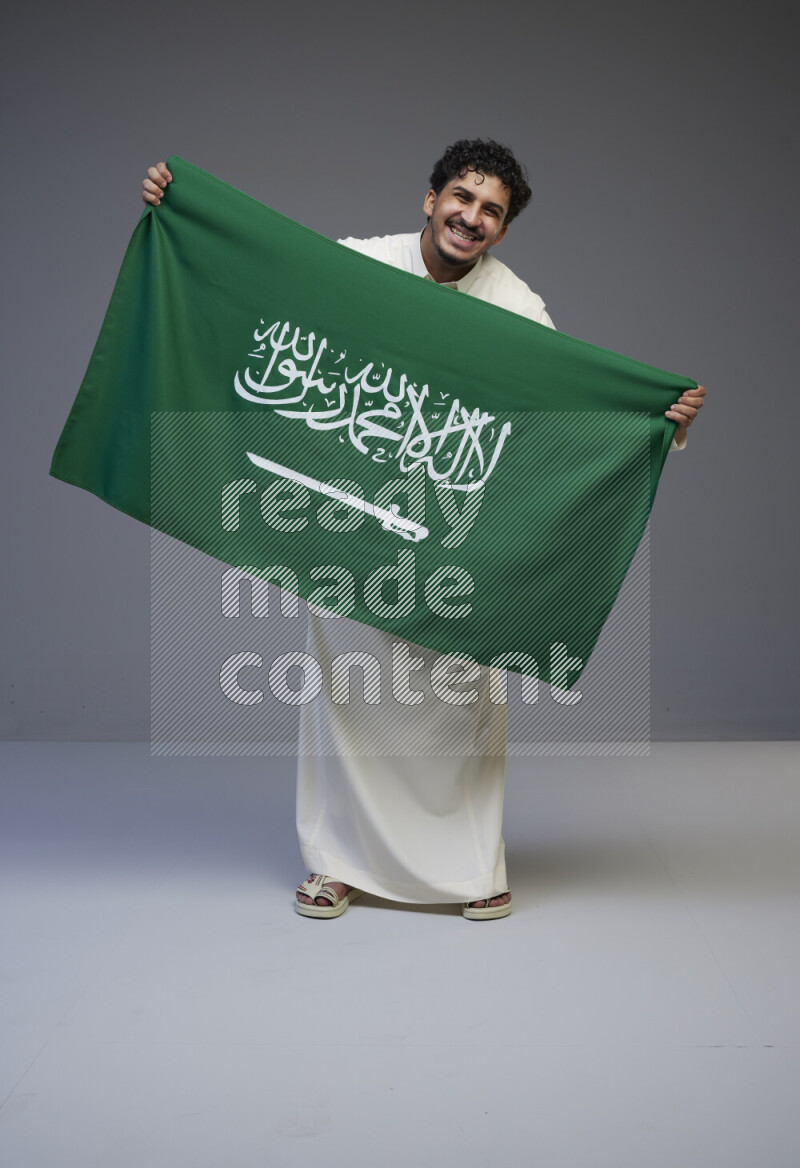 A saudi man standing wearing thob holding big saudi flag on gray background