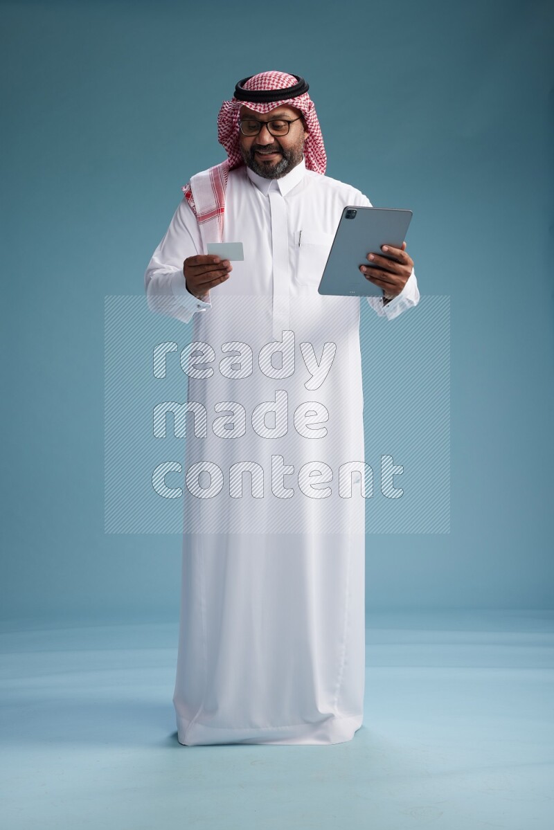 Saudi Man with shimag Standing holding ATM card while working on tablet on blue background