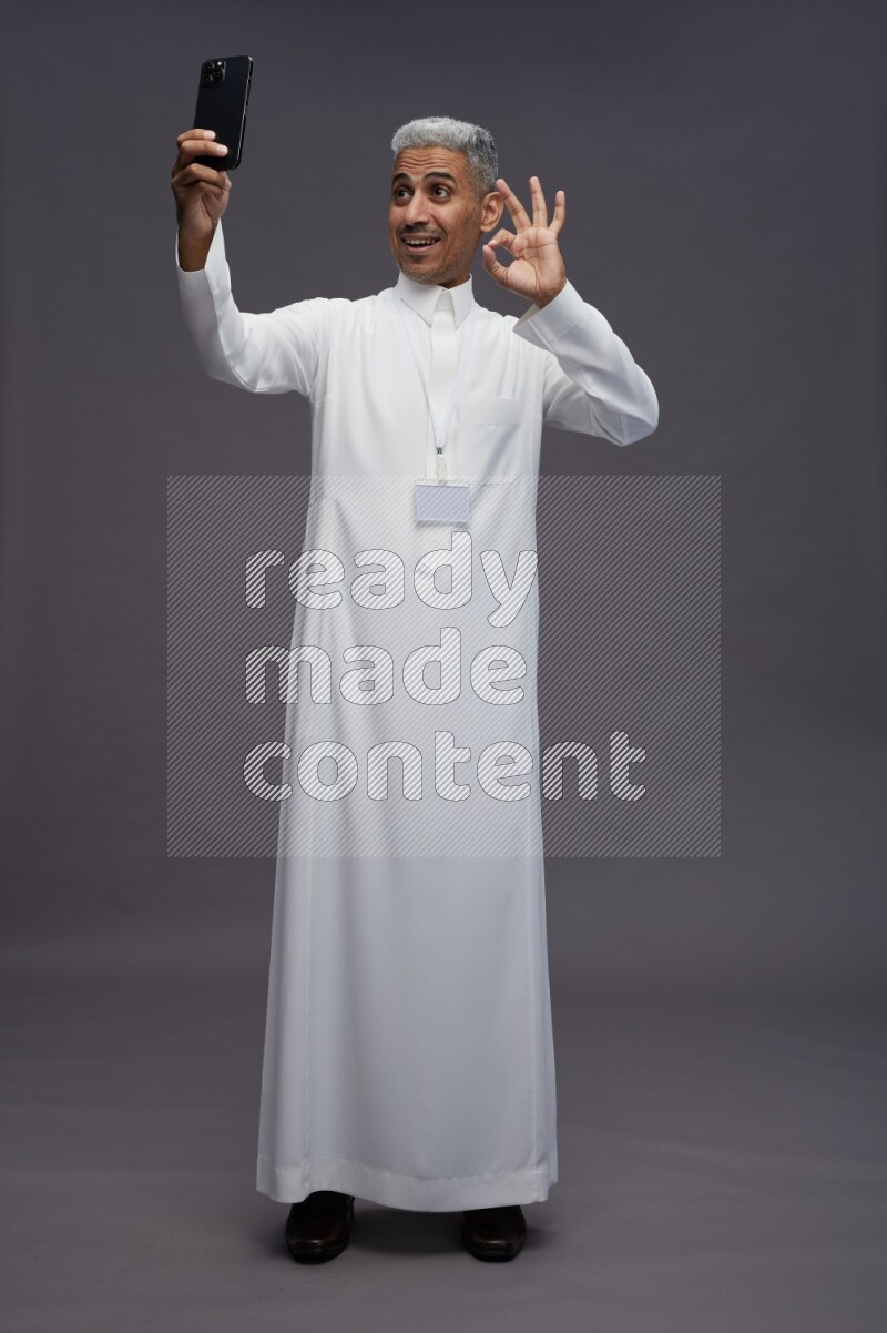 Saudi man wearing thob with neck strap employee badge standing taking selife on gray background