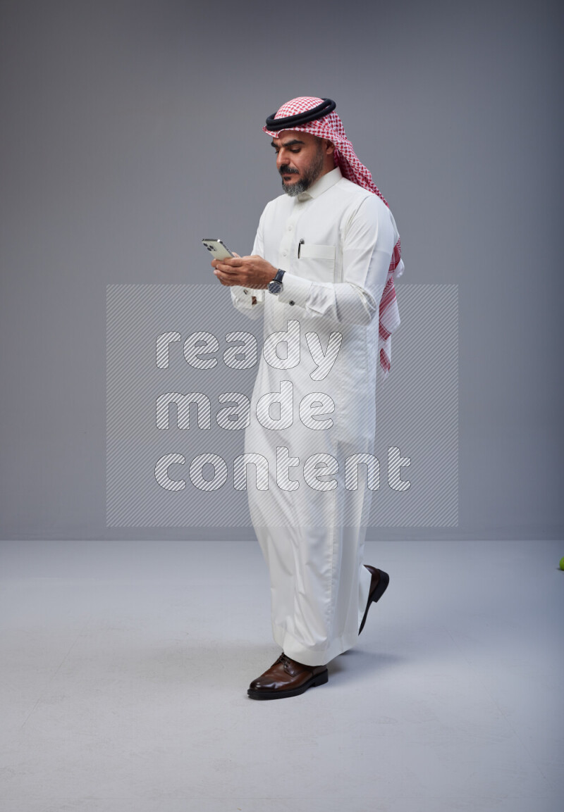 Saudi man Wearing Thob and red Shomag standing texting on phone on Gray background