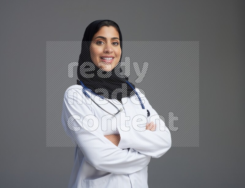 Saudi woman wearing lab coat with stethoscope standing with crossed arms on Gray background