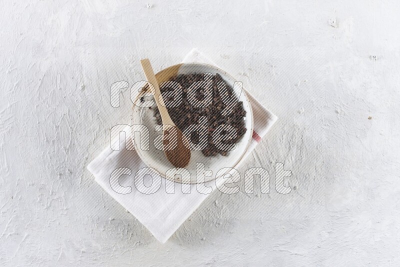 A Pottery plate full of whole cloves and a wooden spoon full of cloves powder in it on a textured white background