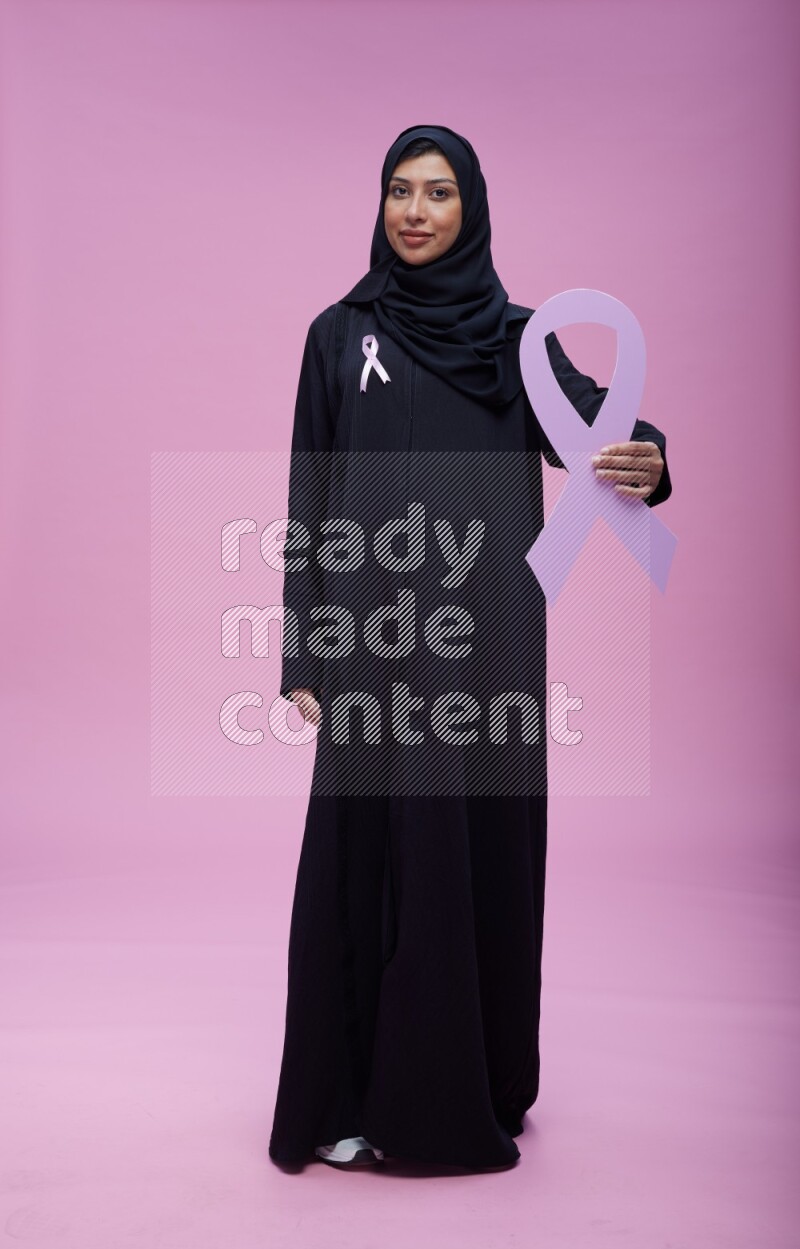 Saudi woman wearing pink ribbon on abaya standing holding awareness ribbon on plain pink background