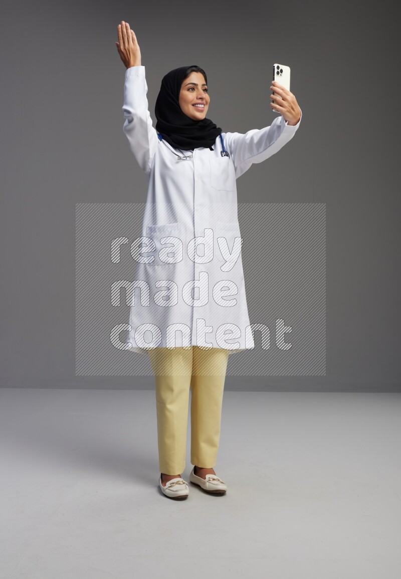 Saudi woman wearing lab coat with stethoscope standing taking selfie on Gray background