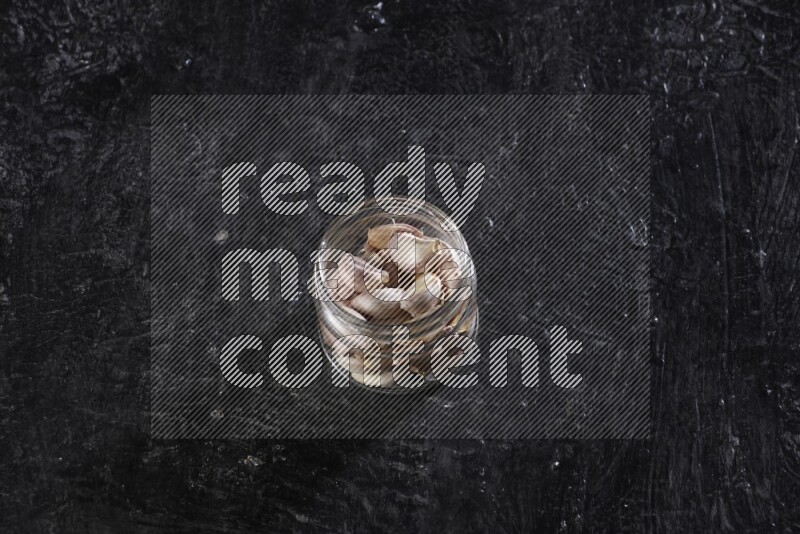 A glass jar full of garlic cloves on a textured black flooring in different angles