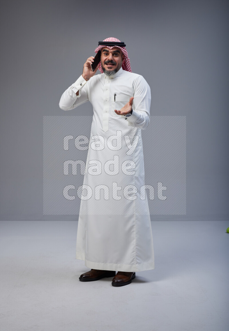 Saudi man Wearing Thob and red Shomag standing talking on phone on Gray background