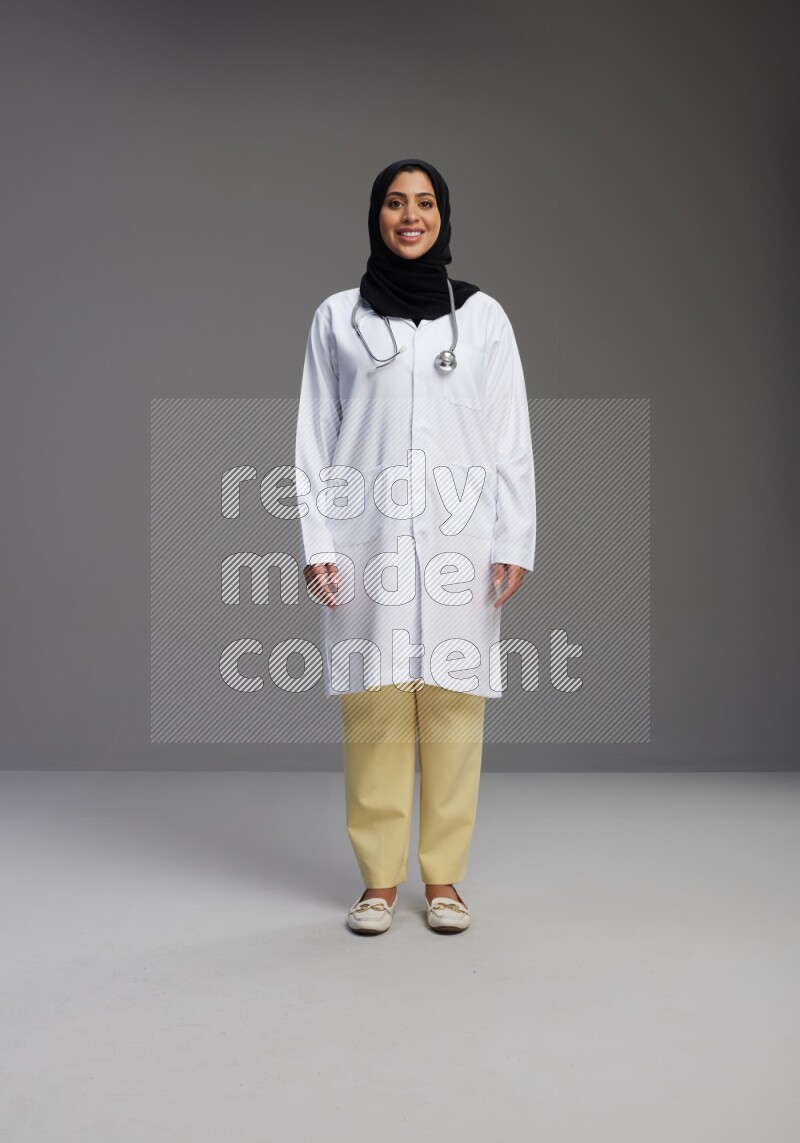 Saudi woman wearing lab coat with stethoscope standing interacting with the camera on Gray background