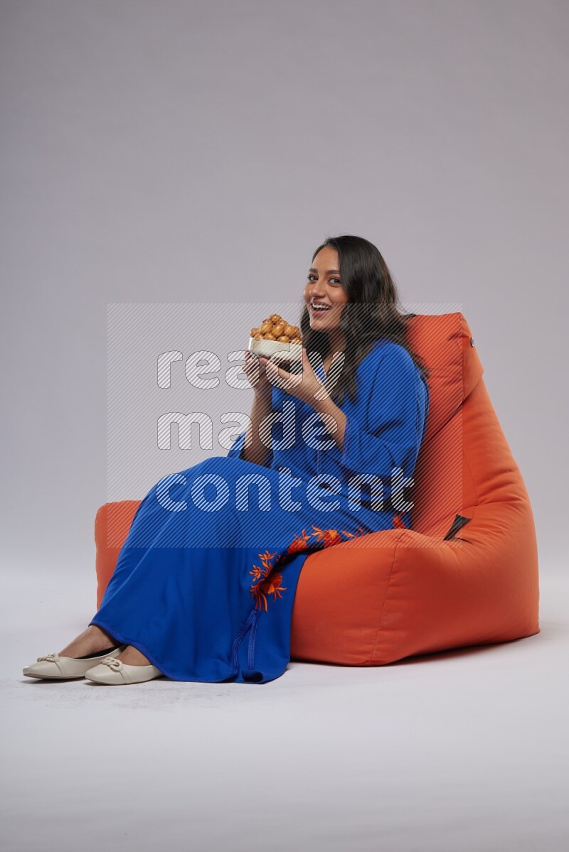 A Woman sitting on an orange beanbag wearing Jalabeya holding a plate of luqaimat