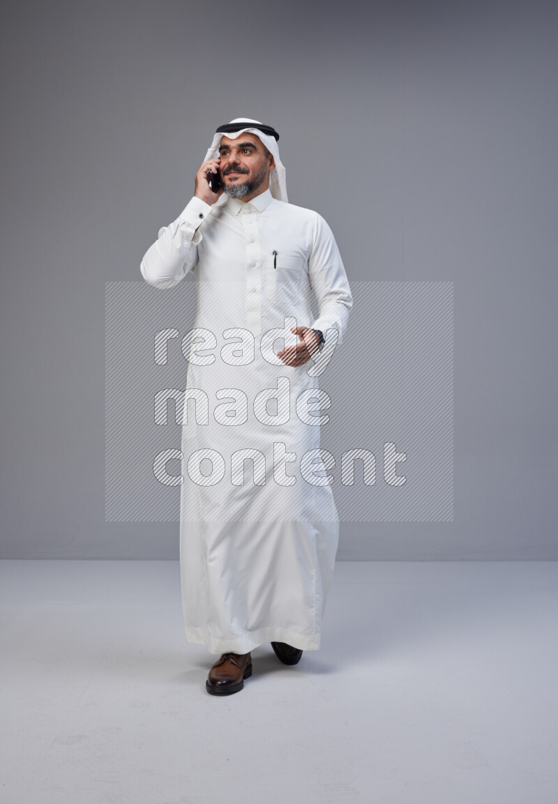 Saudi man Wearing Thob and white Shomag standing talking on phone on Gray background