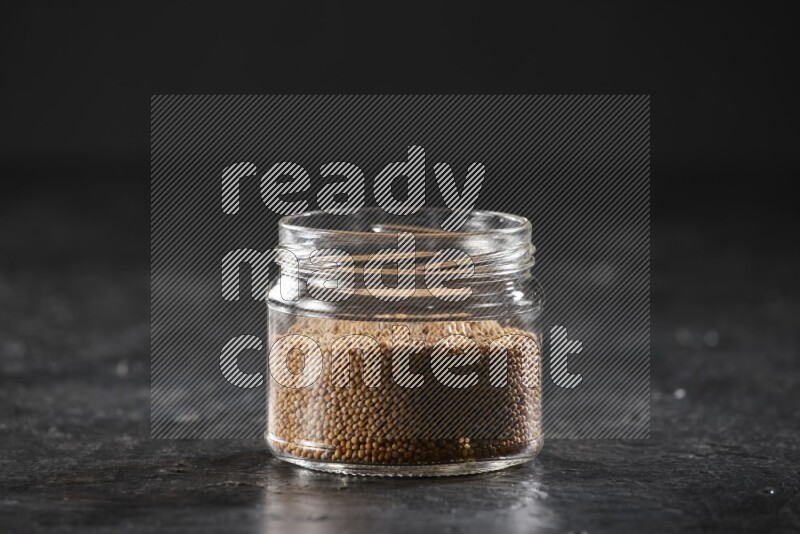 A glass jar full of mustard seeds on a textured black flooring