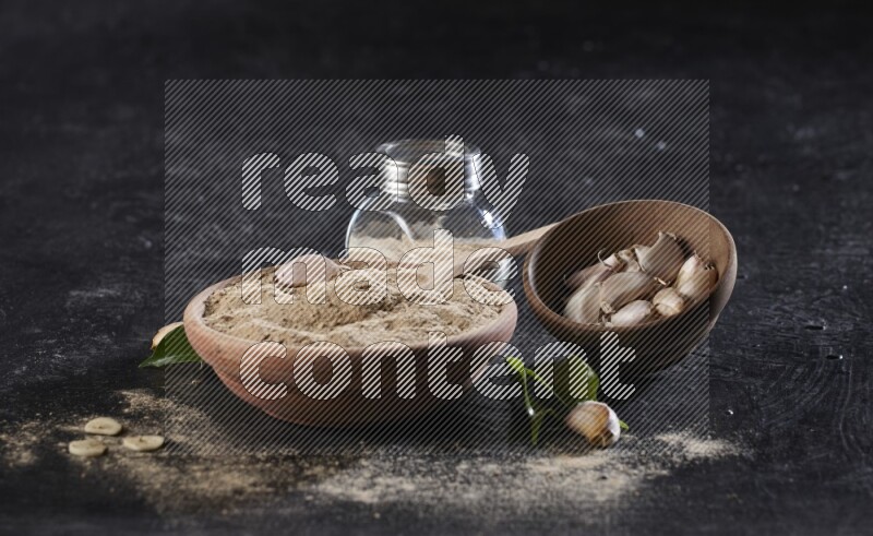 A wooden bowl with a spoon in it and glass spice jar all, full of garlic powder and a wooden bowl full of garlic cloves on a black flooring