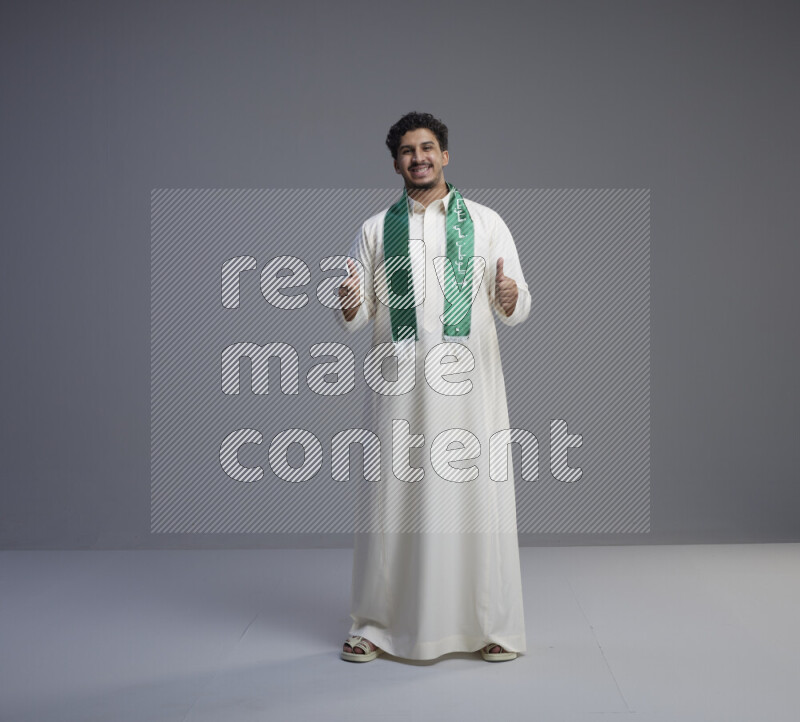 A Saudi man standing wearing thob and Saudi flag scarf on gray background