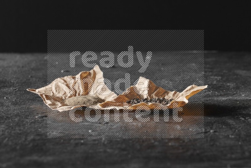 Black pepper and black pepper powder on 2 crumpled paper on a textured black flooring