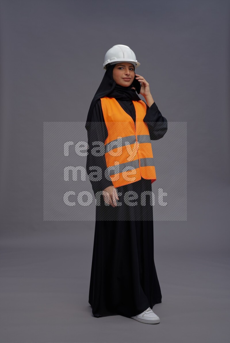 Saudi woman wearing Abaya with engineer vest standing talking on phone on gray background