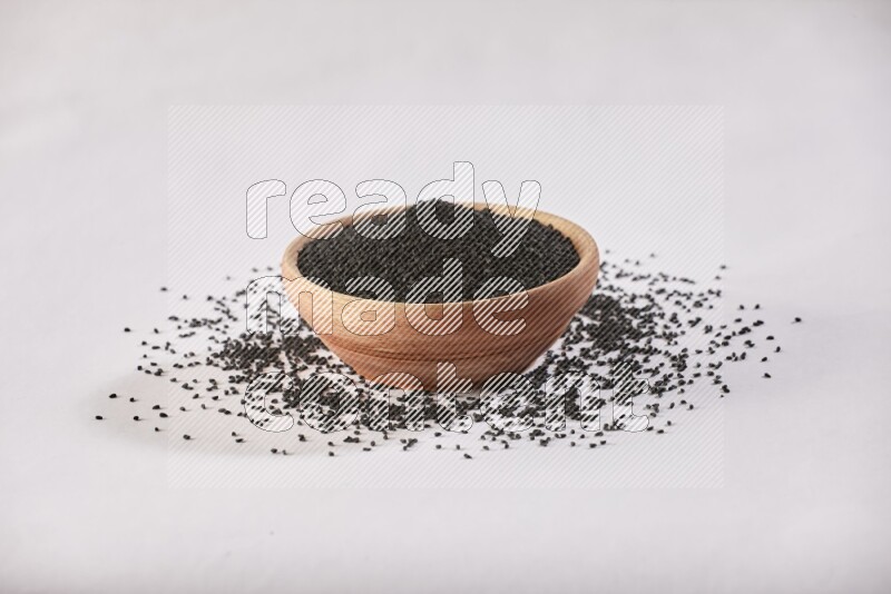 A wooden bowl full of black seeds and more seeds spread on a white flooring