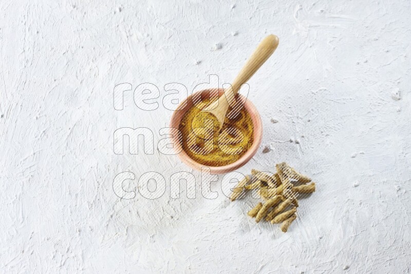 A wooden bowl and wooden spoon full of turmeric powder with dried turmeric fingers beside it on textured white flooring