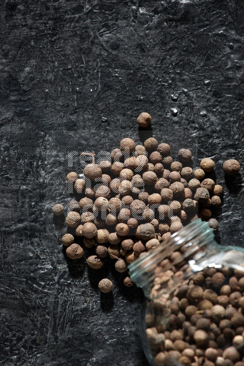 A flipped glass spice jar full of allspice whole balls and the balls spilled out of it on a textured black flooring