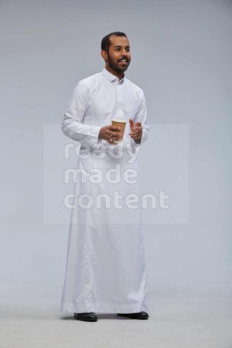 Saudi man wearing thob standing holding paper cup on gray background