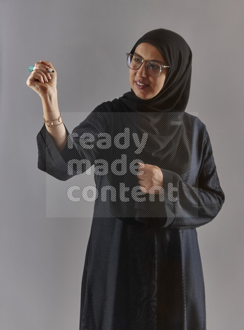 Woman in black abaya, hijab and glasses holding a marker pen to write on a transparent board with different reactions and expressions on grey background