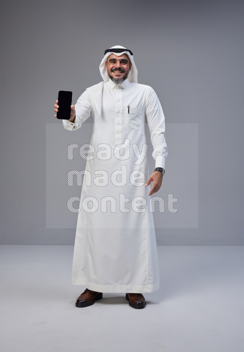 Saudi man Wearing Thob and white Shomag standing showing phone to camera on Gray background