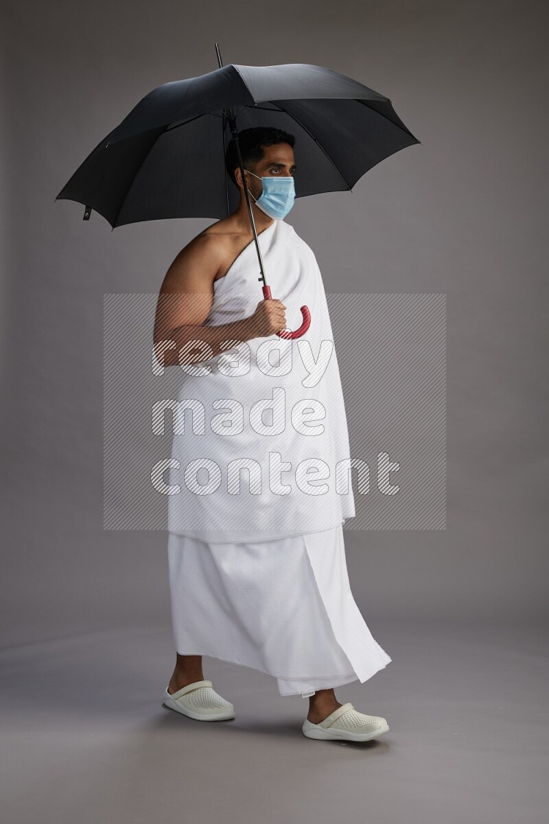 A man wearing Ehram and mask Standing holding umbrella on gray background on gray background
