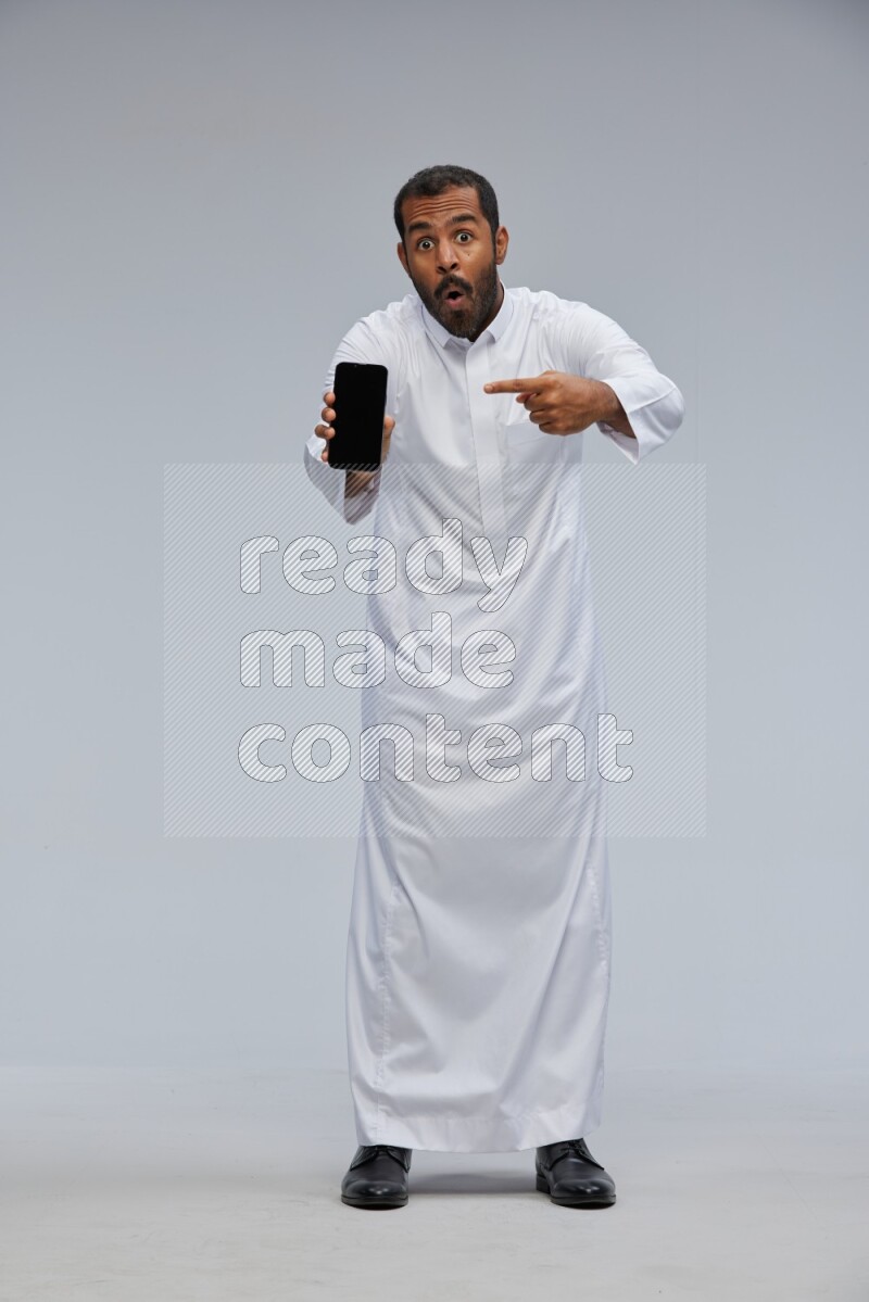 Saudi man wearing Thob standing showing phone to camera on Gray background