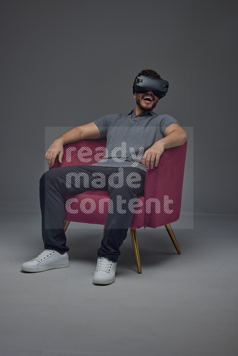 A man wearing casual and wearing VR while sitting on a burgundy chair eye level on a gray background